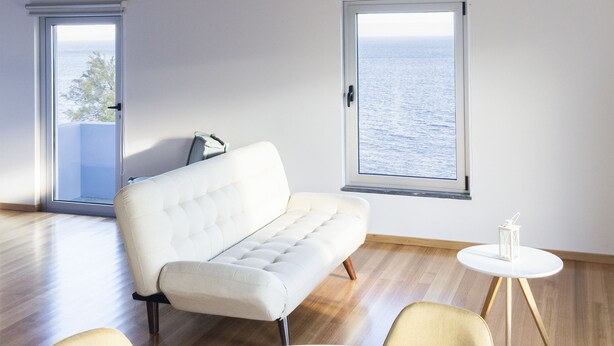 A modern room with wooden beams, a white sofa, a round table, and a view of the sea through a window. - Lofts Azul Pastel (Horta)