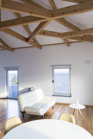 A modern room with wooden beams, a white sofa, a round table, and a view of the sea through a window.