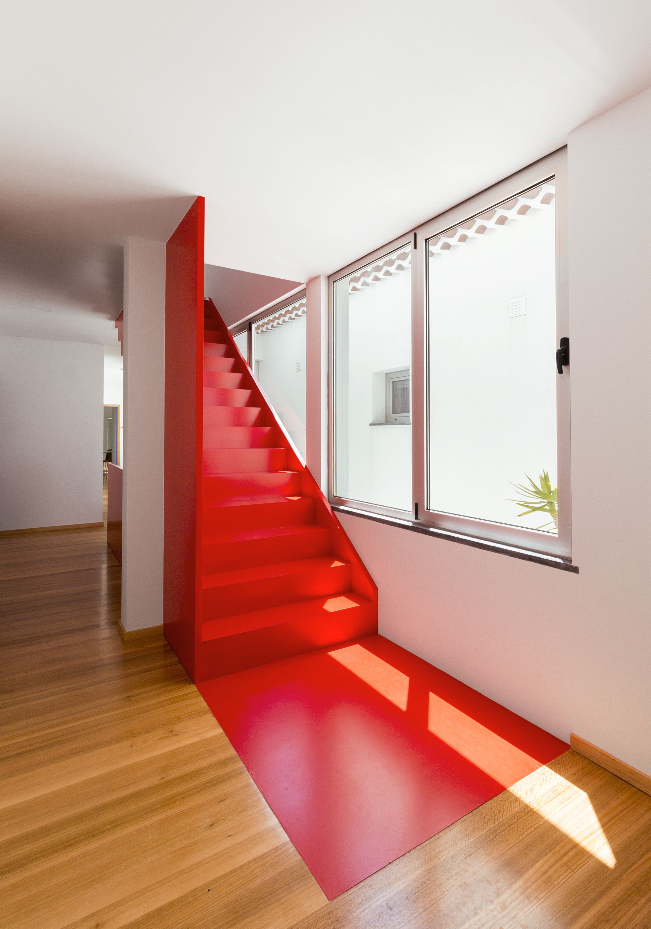 A modern interior with a wooden floor, a red staircase, and large windows.