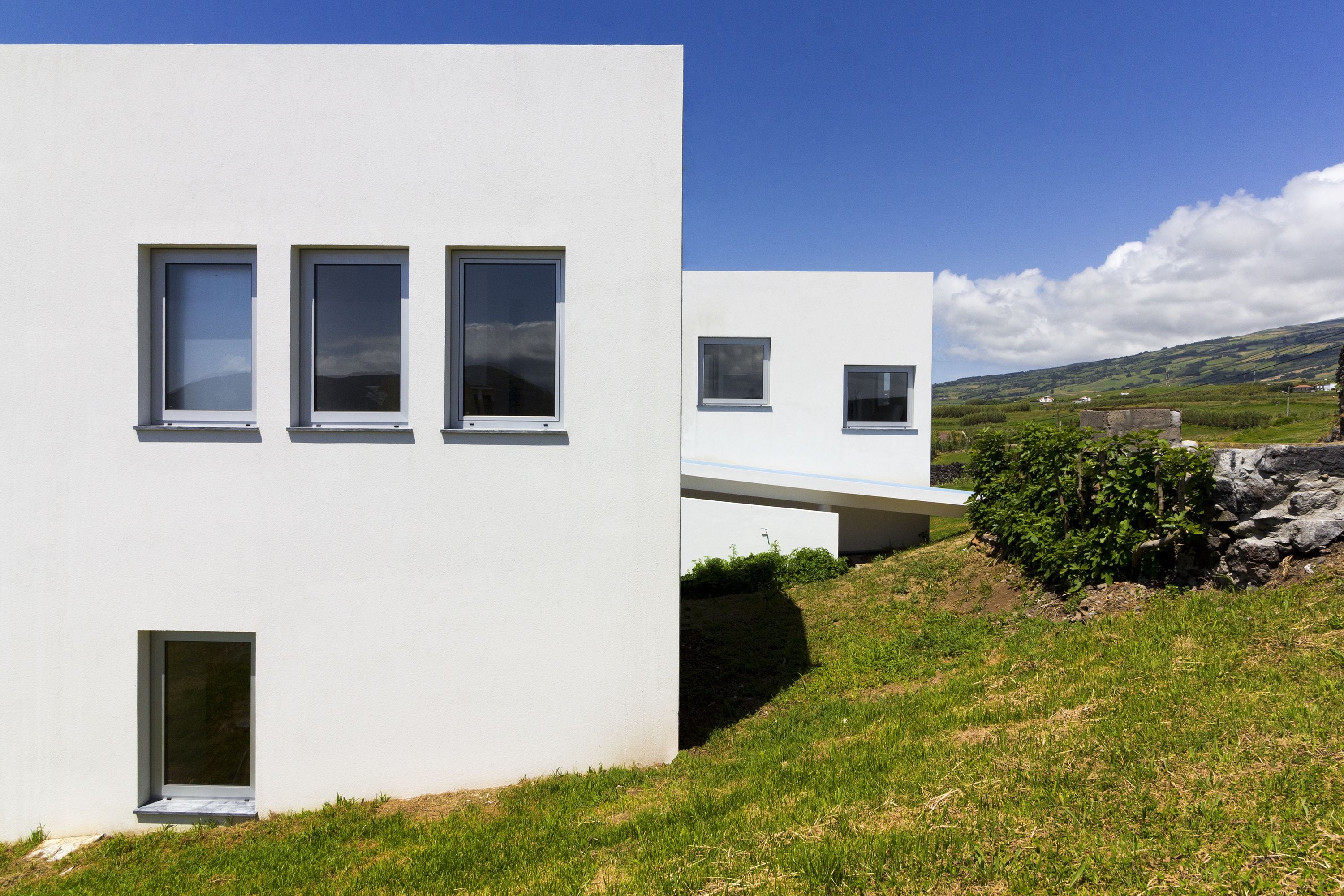 A modern building with three windows on the facade, a grassy area in front, and a stone wall to the right.