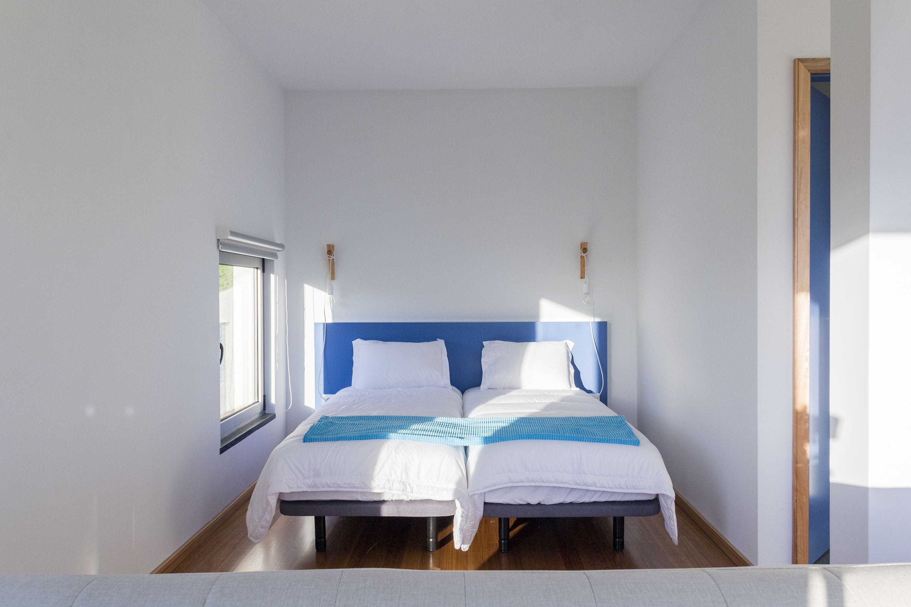 A double bed with a blue headboard in a bright room with a window and a door.