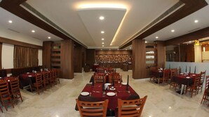 A spacious dining area with multiple tables and chairs, a central chandelier, and a decorative wall in the background. - Rathna Residency (Salem)