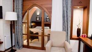 Une chambre d’hôtel dotée d’une porte à arc en bois donnant sur une chambre à coucher, d’un coin salon avec un fauteuil, d’une table de chevet avec une lampe et d’un vase contenant des branches séchées.