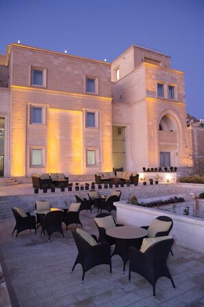 An outdoor seating area with multiple tables and chairs, a stone building with illuminated windows, and a clear sky. - Reyhani Kasri - Special Class (Mardin)