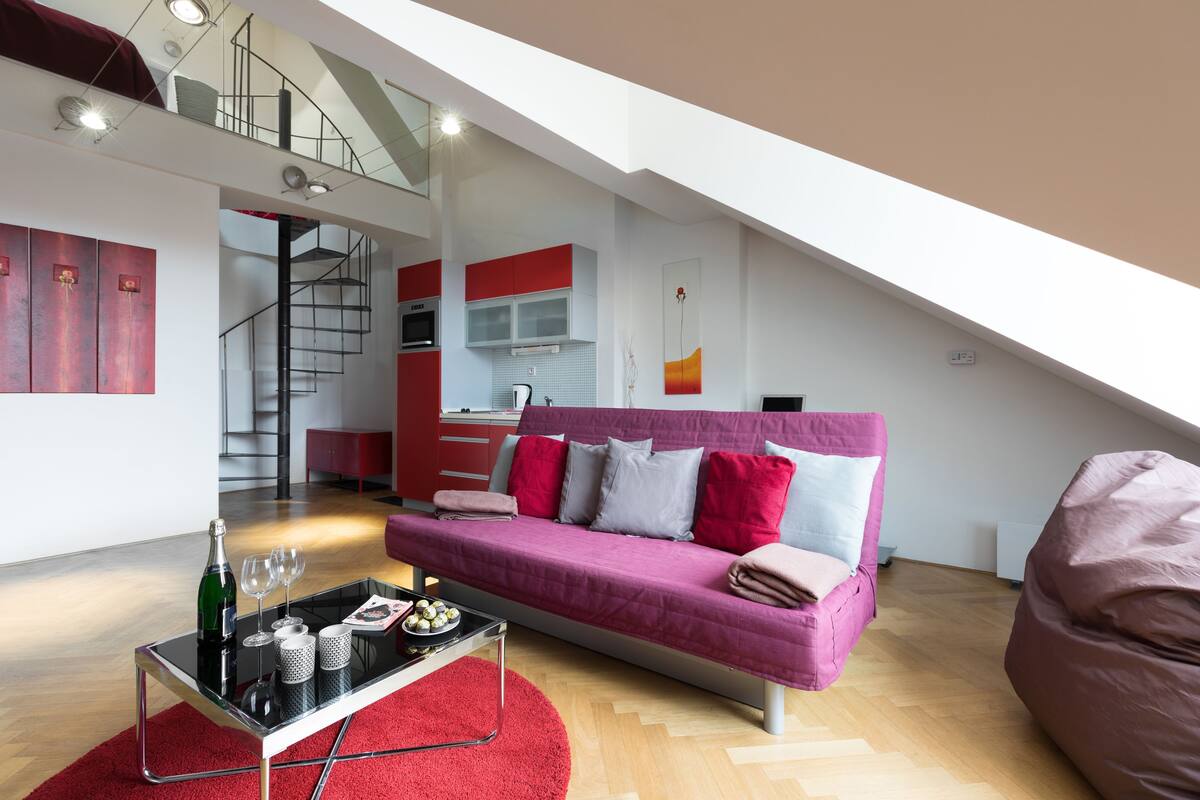 Un loft moderno con cama en altura, sofá rosa, zona de comedor y escalera.