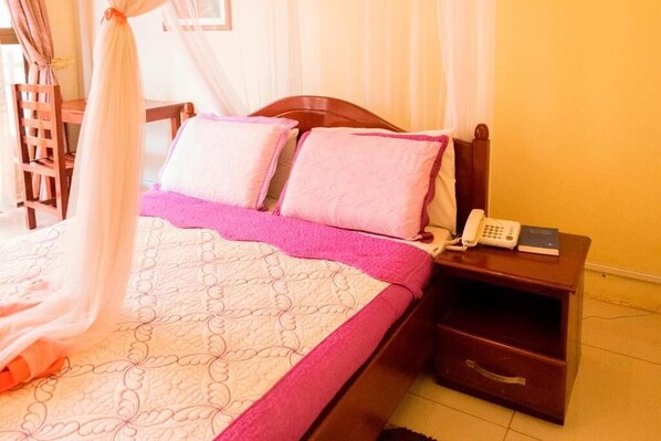A bed with a pink and white quilt, a bedside phone, and a wooden nightstand. - Kahondo Buffalo Hotel (Kampala)
