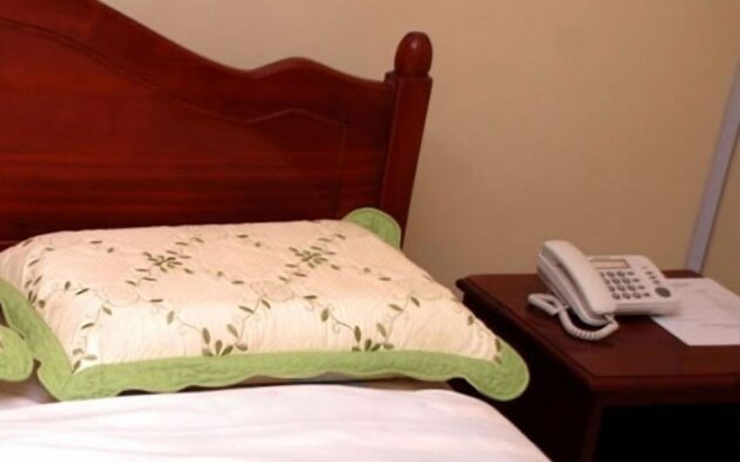 Una cama con un cojín estampado con motivos florales y detalles en verde, un teléfono en la mesita de noche y un cabecero de madera.