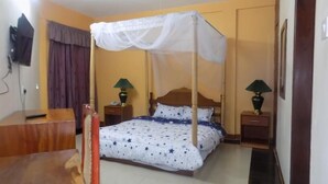 A hotel room with a bed, two bedside tables with lamps, a desk, a chair, a TV mounted on the wall, and a curtain. - Kayegi Hotel (Mbale)