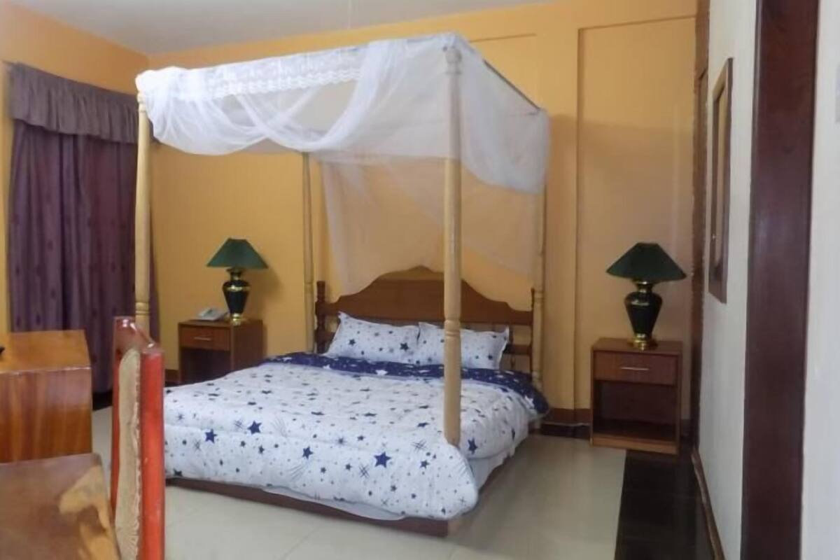 Una habitación de hotel con una cama, dos mesitas de noche con lámparas, un escritorio, una silla, un televisor en la pared y una cortina.