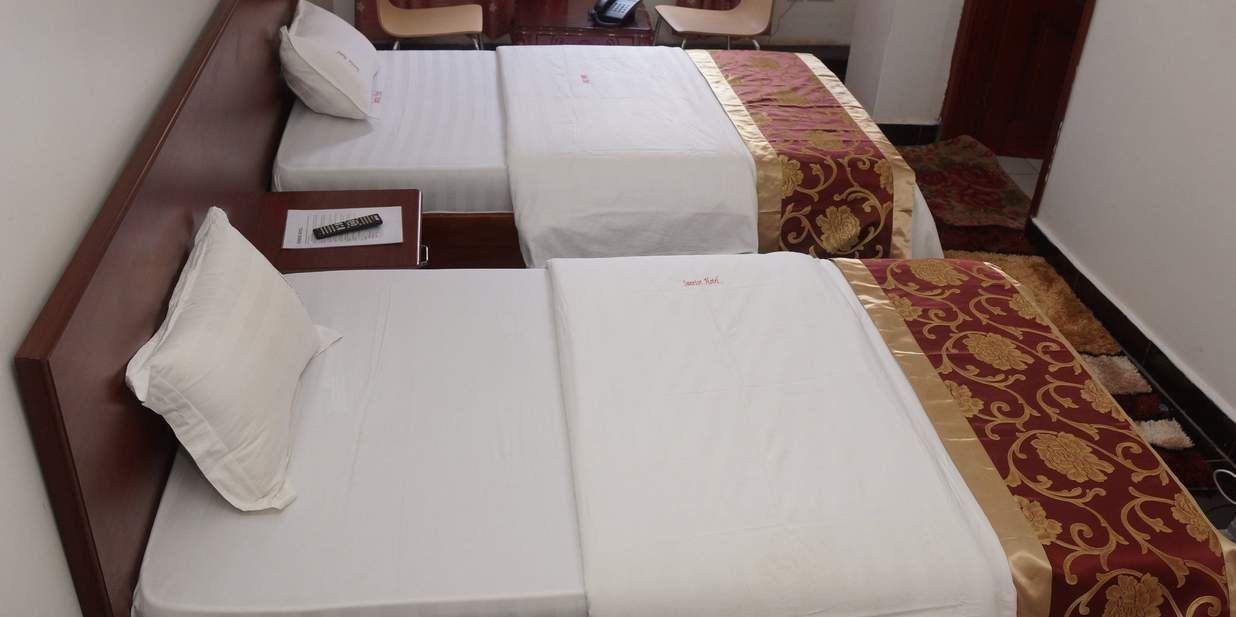 A hotel room with two single beds, a wooden headboard, a bedside table with a remote control, and a patterned carpet.