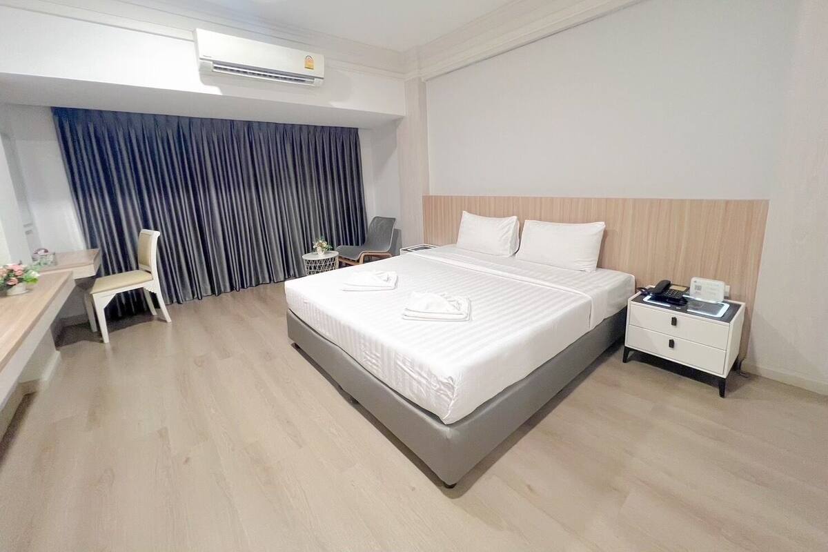A hotel room with a bed, a desk, a chair, a nightstand, and an air conditioning unit.
