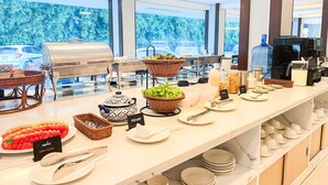 Montaje de bufé con diversos alimentos, utensilios, una máquina de café y vistas a árboles a través de amplias ventanas.