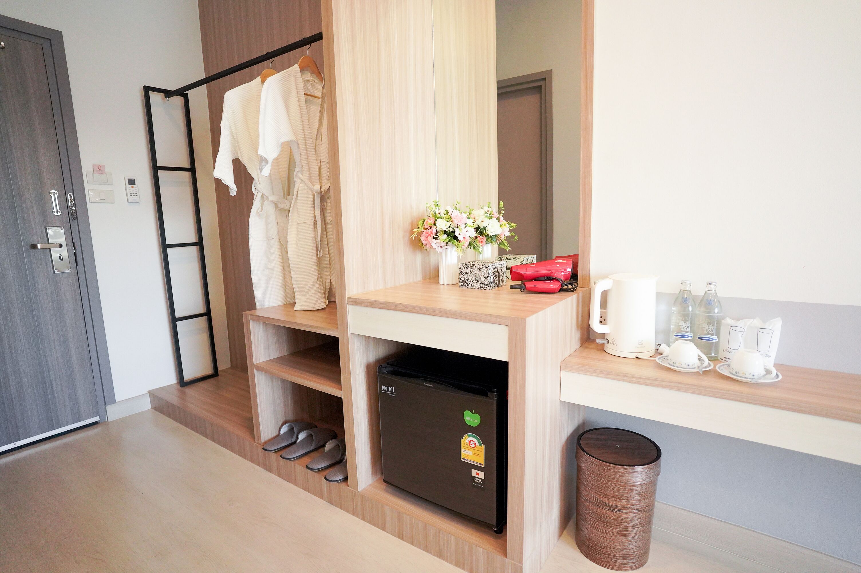 Ein kompaktes Hotelzimmer mit Holzboden, einem kleinen schwarzen Kühlschrank, einem Holzregal mit Schuhen, einem aufgehängten Bademantel, einer kleinen Mülltonne sowie einem Tisch mit Wasserkocher, Tassen und einer Blumenvase.