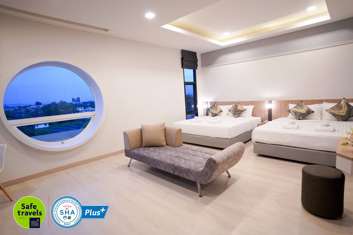 A modern hotel room with a large round window, two beds, a sofa, and a small ottoman.