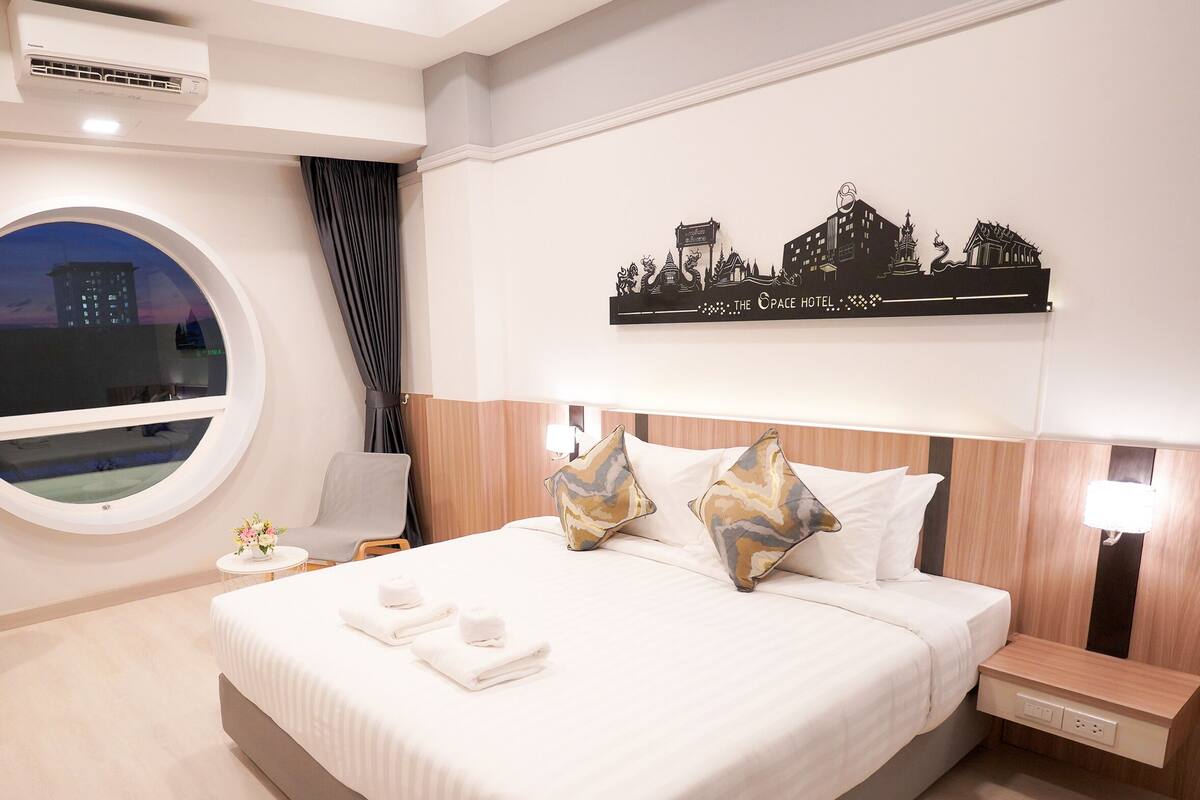 A hotel room with a large bed, a round window, a cityscape wall art, a nightstand, and a small table with a vase of flowers.
