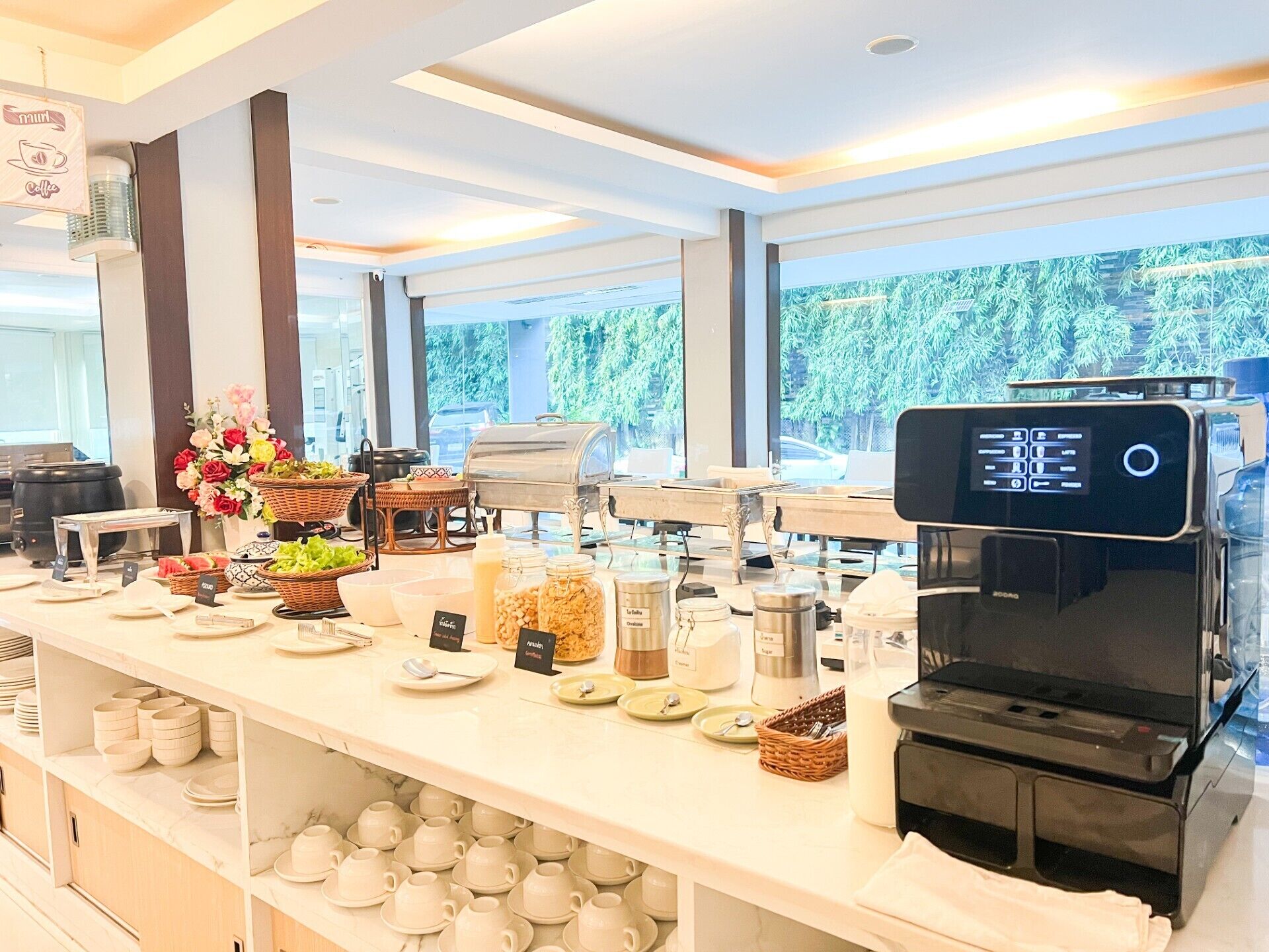 A buffet setup with various food items, a coffee machine, and a view of greenery outside.