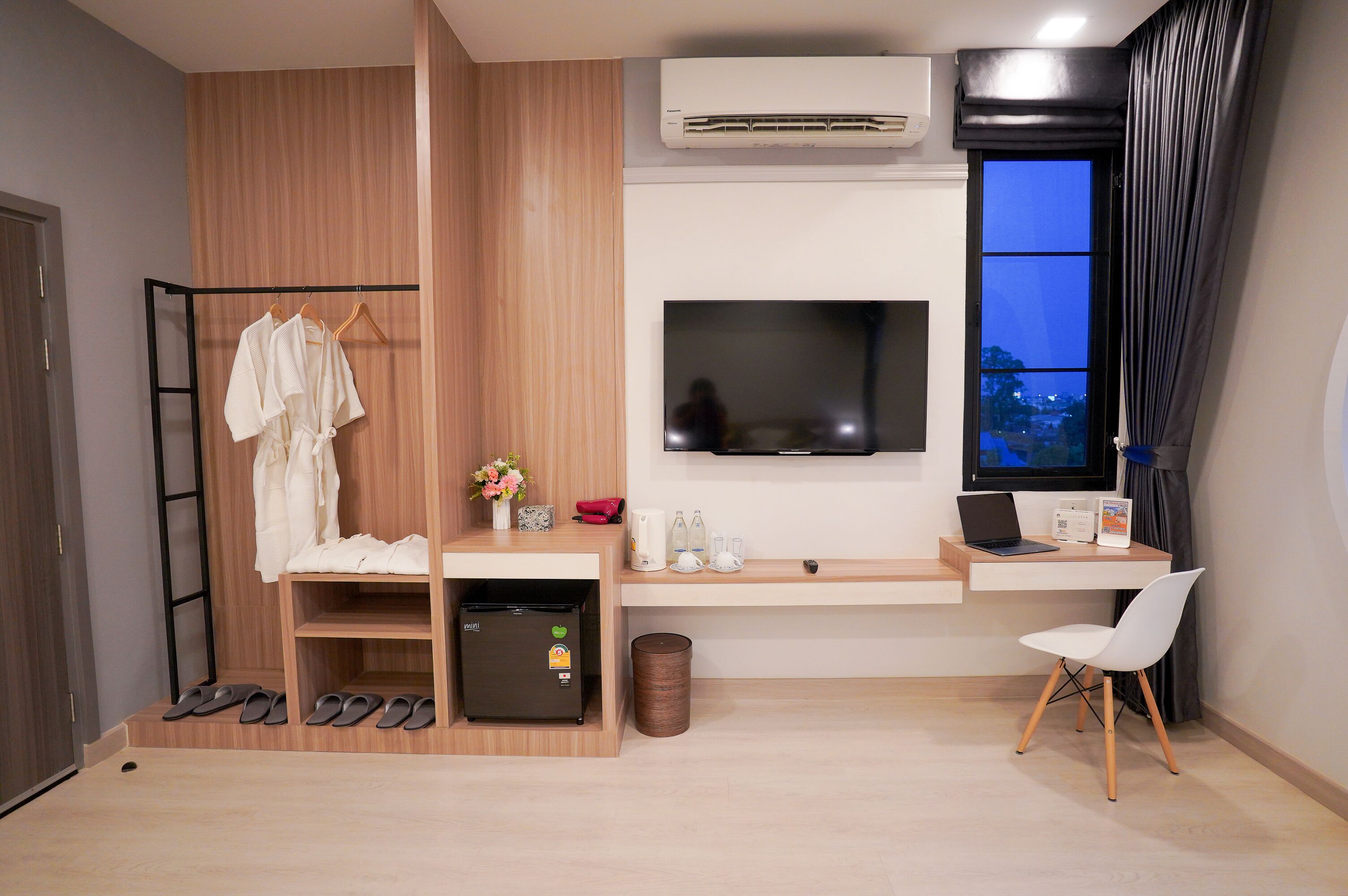 A modern room with a wooden wardrobe, a small fridge, a TV mounted on the wall, a desk with a laptop, and a chair.