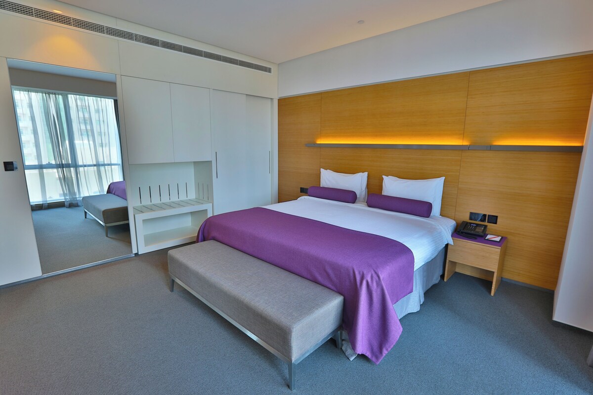 A modern hotel room with a large bed, a bench, a nightstand, a wardrobe, and a mirror.