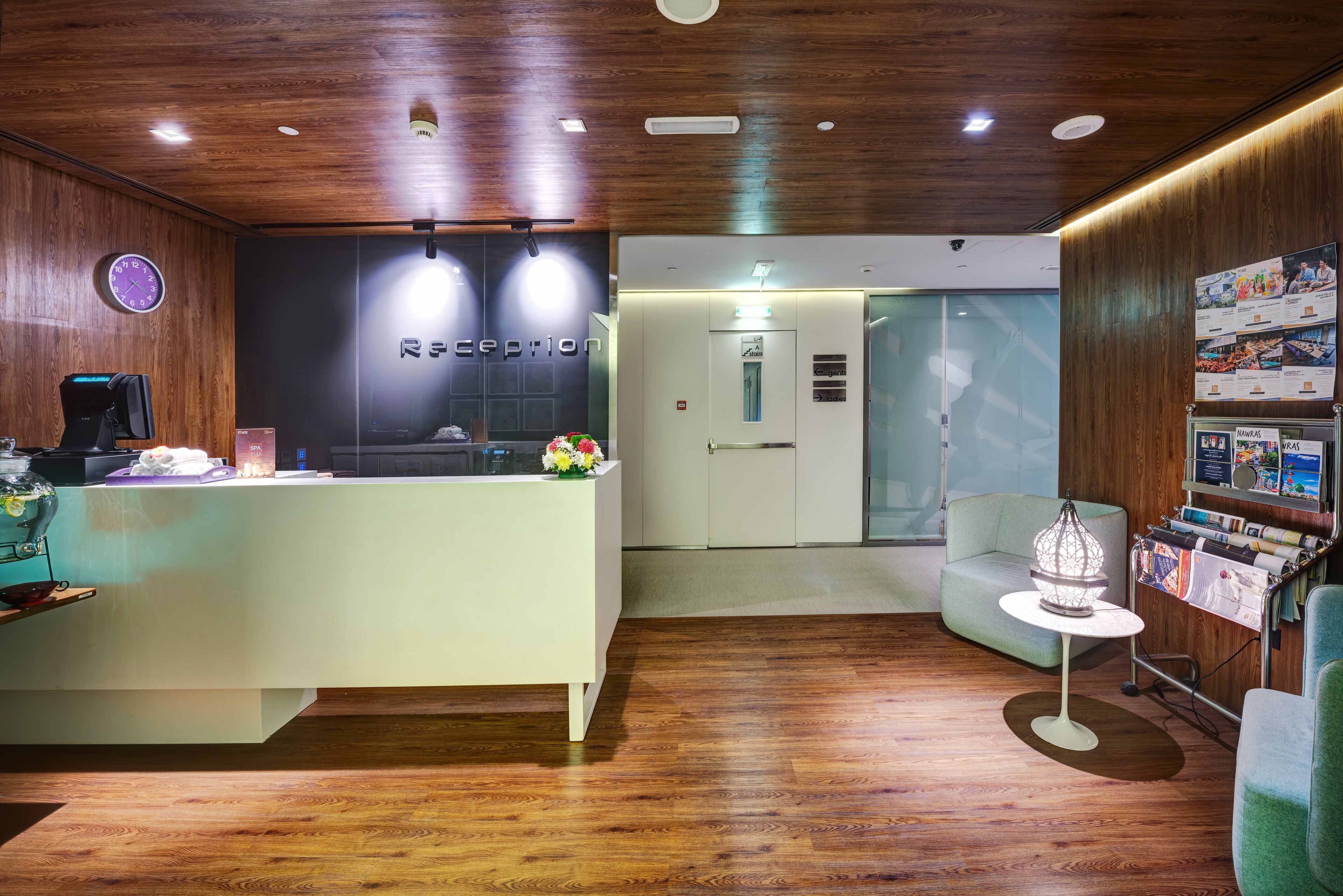 Un área de recepción con techo de madera, mostrador de recepción, reloj, dispensador de agua, zona de estar y un atril para revistas.