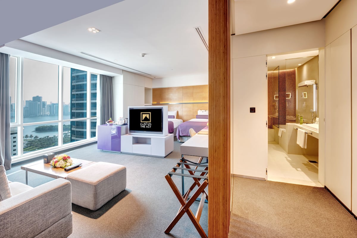 A modern hotel room with a large window, a flat-screen TV, a sofa, a coffee table, and a bathroom visible through an open door.