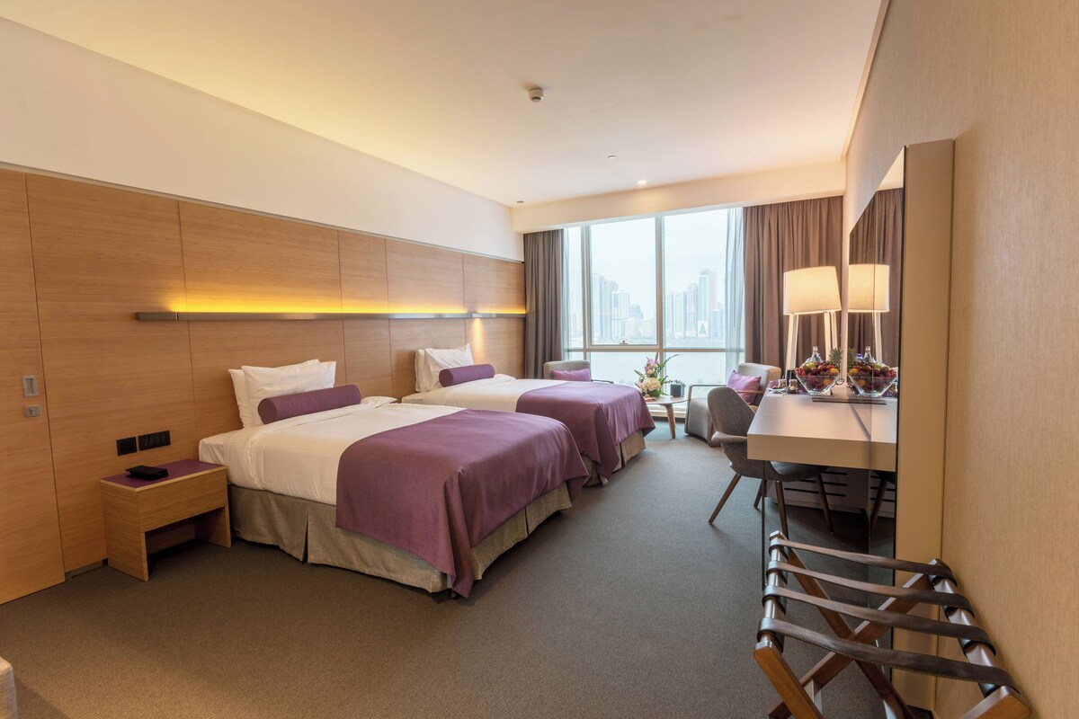A hotel room with two beds, a desk with a lamp, a chair, and a view of the city.