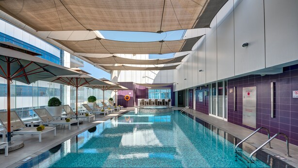 An indoor swimming pool with sun umbrellas, lounge chairs, and a view of the cityscape. - The Act Hotel Sharjah (Sharjah)