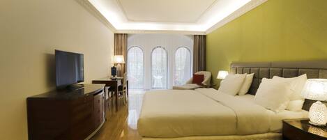 A hotel room with a large bed, a TV, a desk, and a view of the outdoors through arched windows.