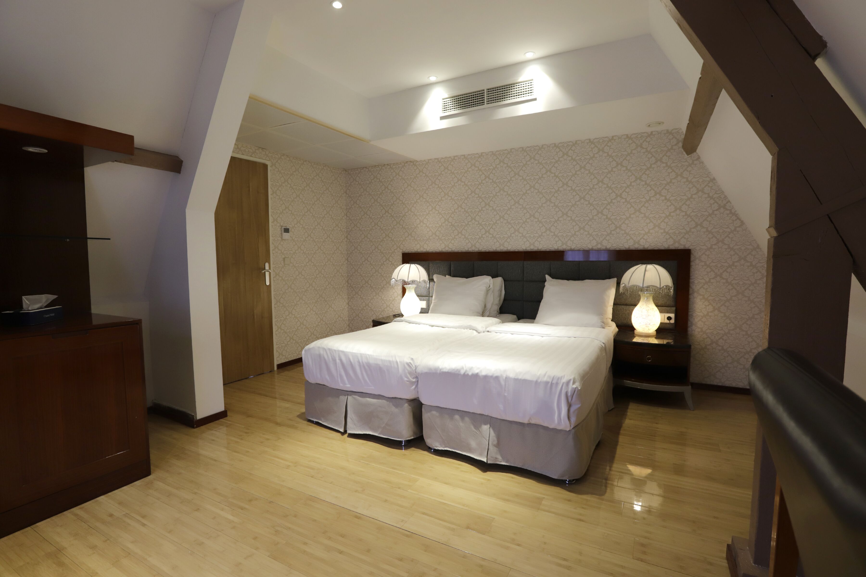 Un dormitorio con una cama grande, una mesita de noche con lámpara, un aparador de madera y una puerta que da a otra habitación.