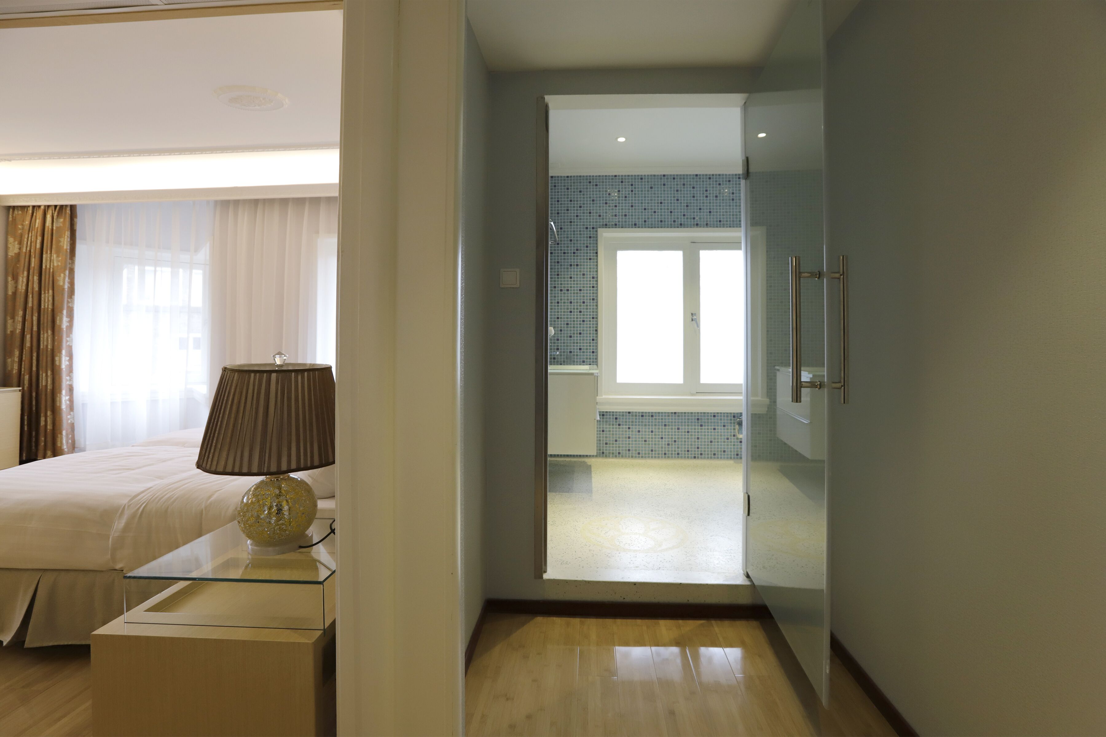 A modern hotel room with a large bed, a bedside table with a lamp, and a view of the bathroom through an open door.