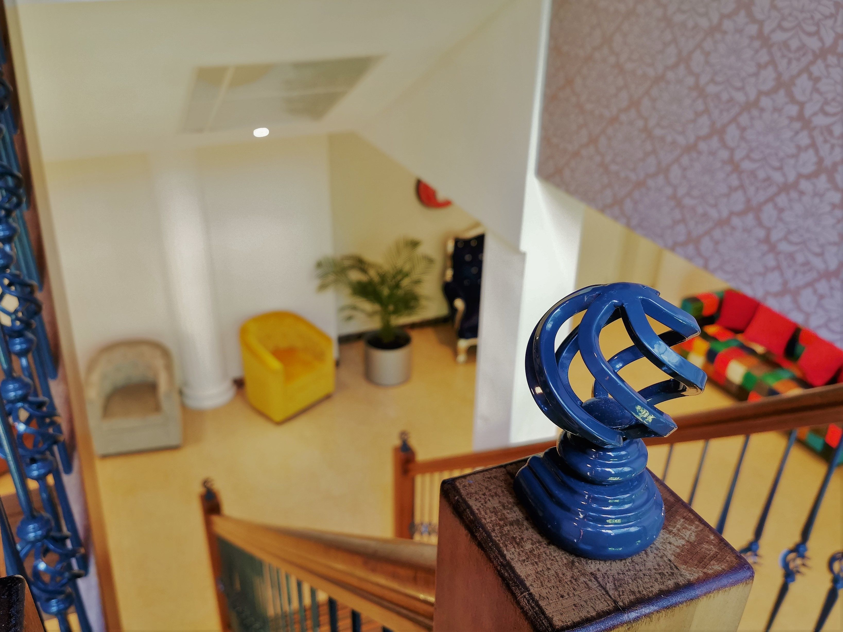 Una escalera con barandilla de metal azul, una silla amarilla, una planta en maceta y un sofá estampado en rojo y verde al fondo.