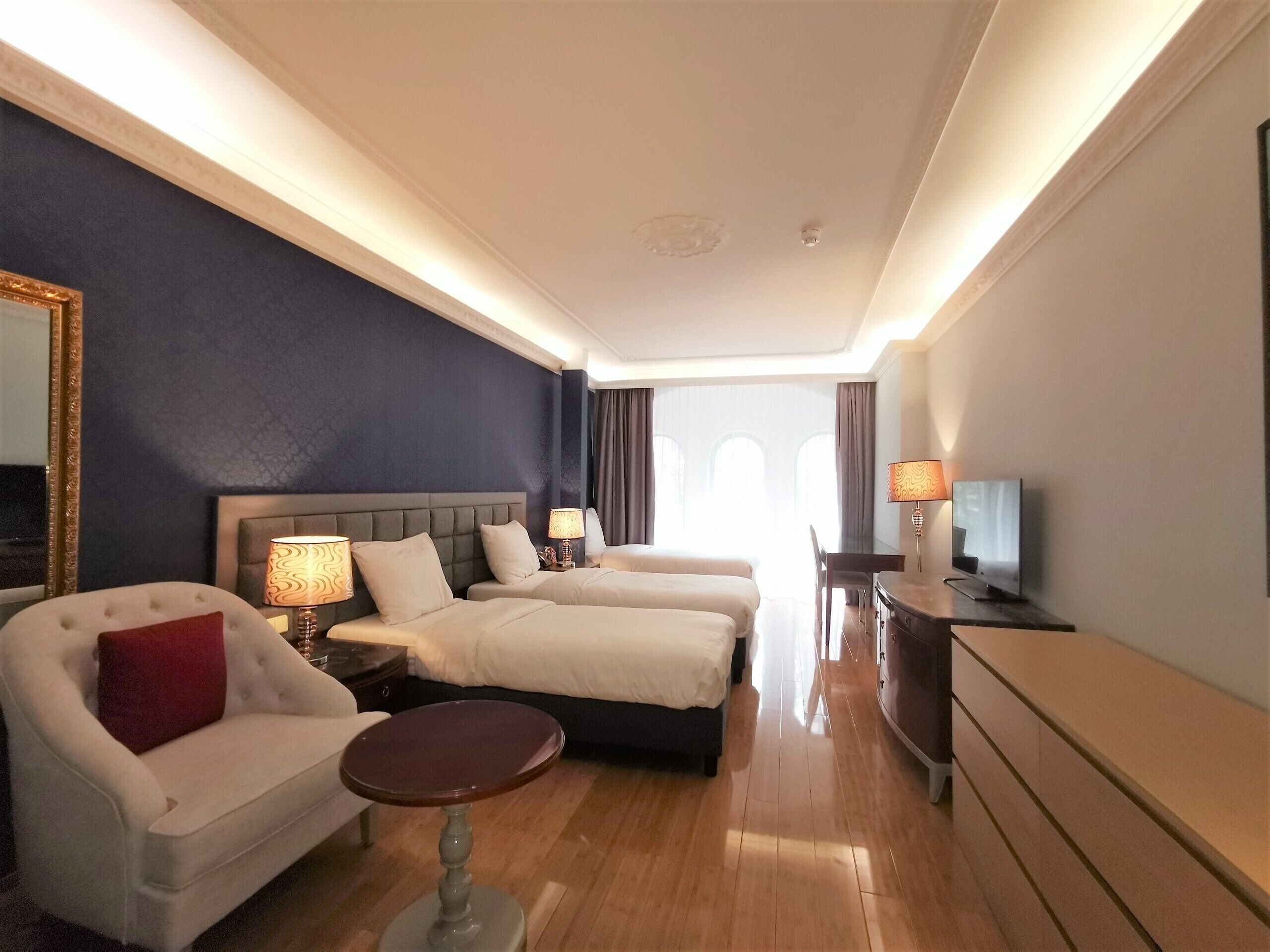 A hotel room with a bed, two bedside tables with lamps, a chair, a small round table, a TV, and a dresser.
