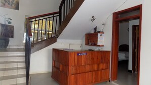 Mostrador de recepción con la etiqueta "RECEPTION" en el vestíbulo de un hotel, con una escalera y una cama a lo lejos.
