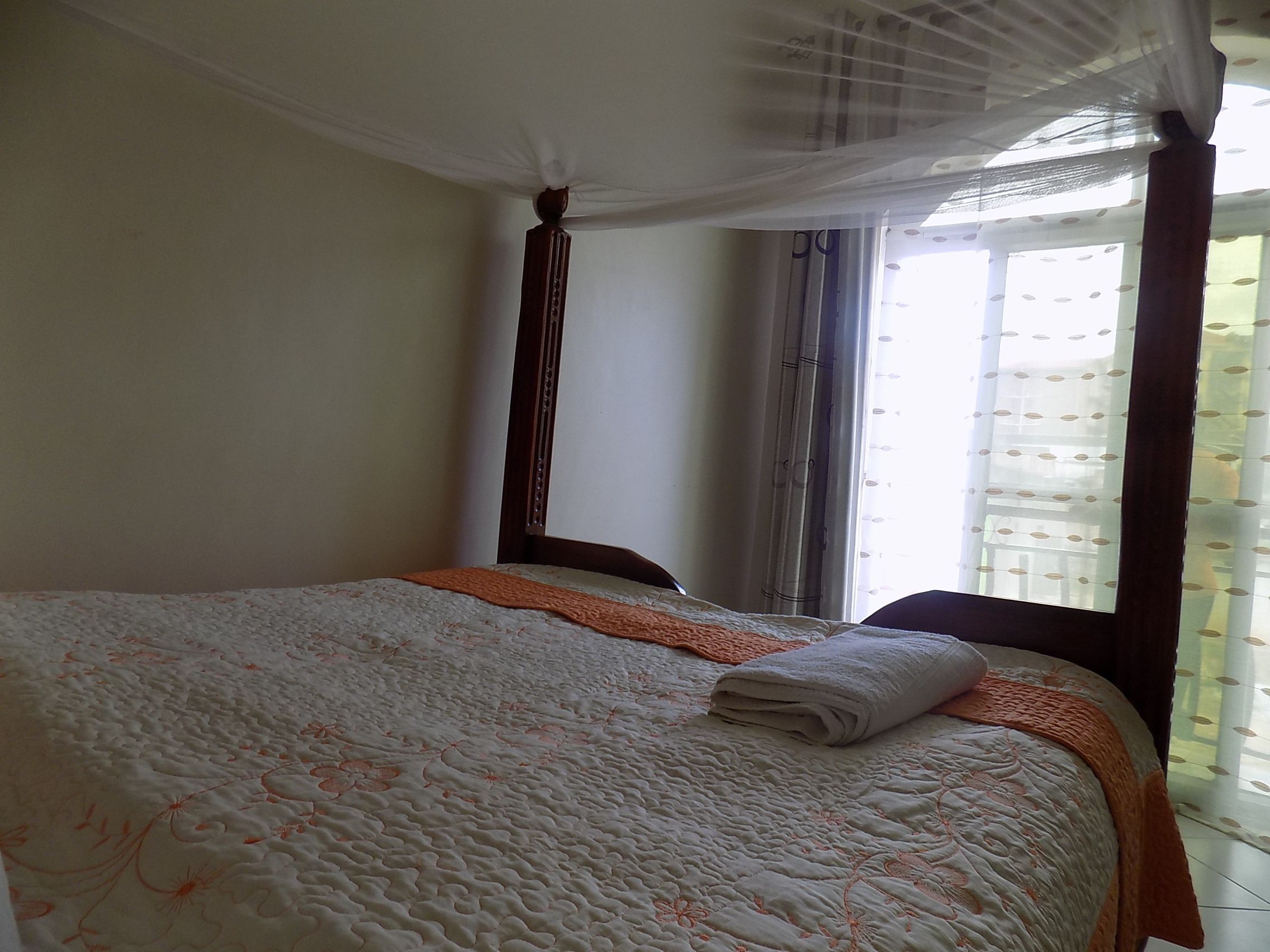 Un lit bien fait, avec une couette blanche et des touches de couleur orange, une fenêtre ornée de rideaux légers et une tête de lit en bois.