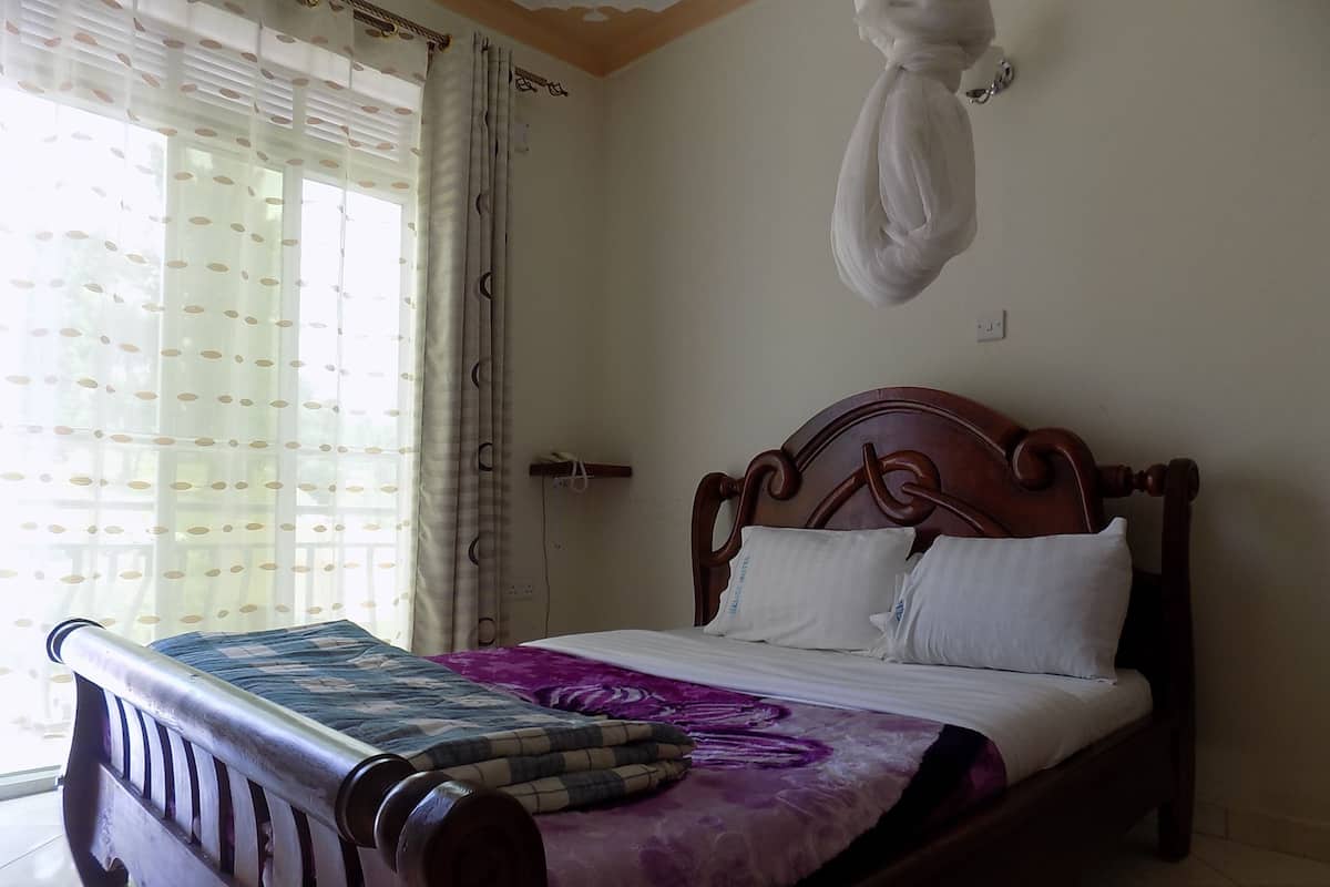 Un dormitorio con cama de madera, colcha morada y azul, almohadas blancas y una ventana con cortinas translúcidas.