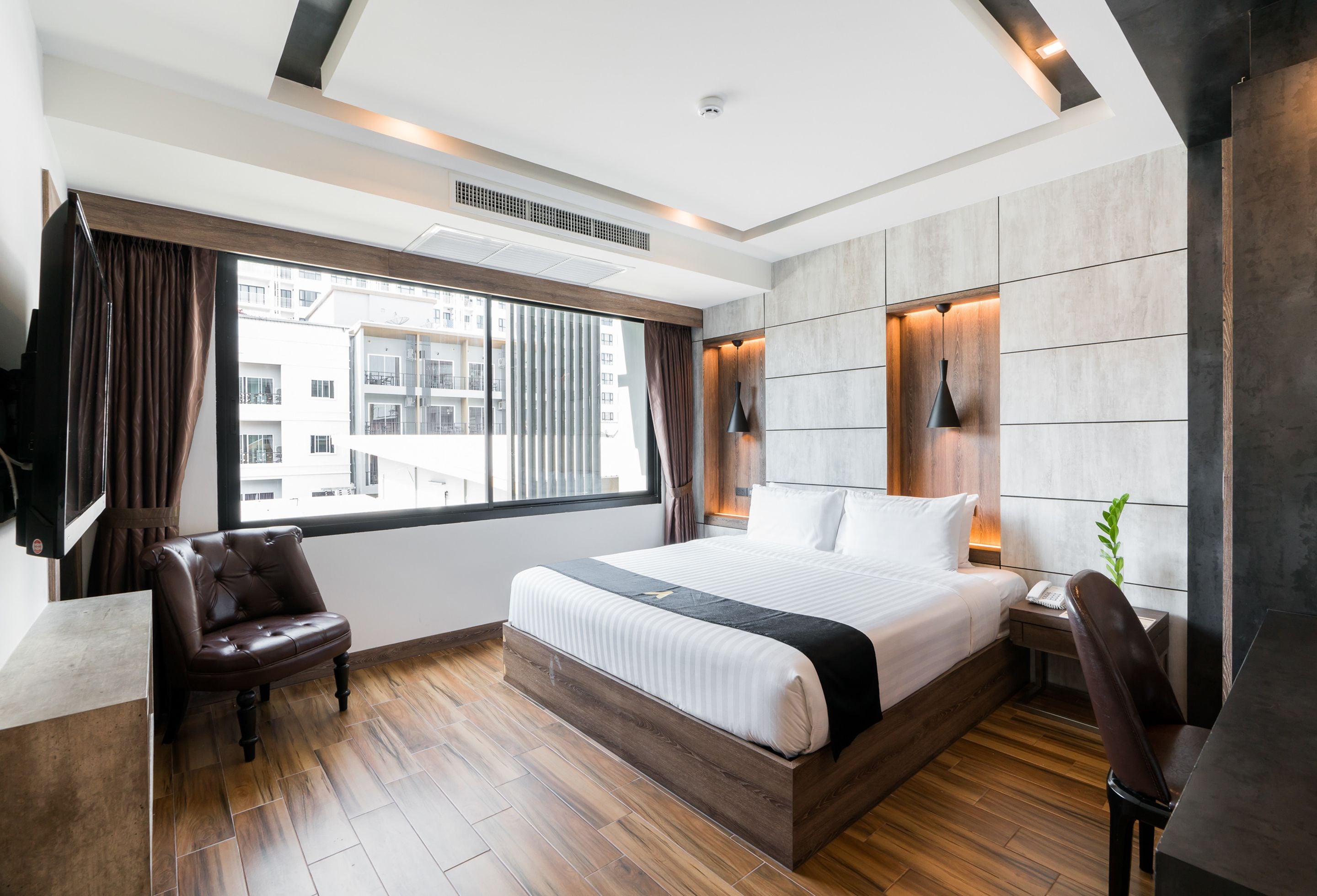 Quarto de hotel moderno com uma cama grande, uma escrivaninha e uma cadeira. Há uma janela com cortinas, uma planta e vista para edifícios.