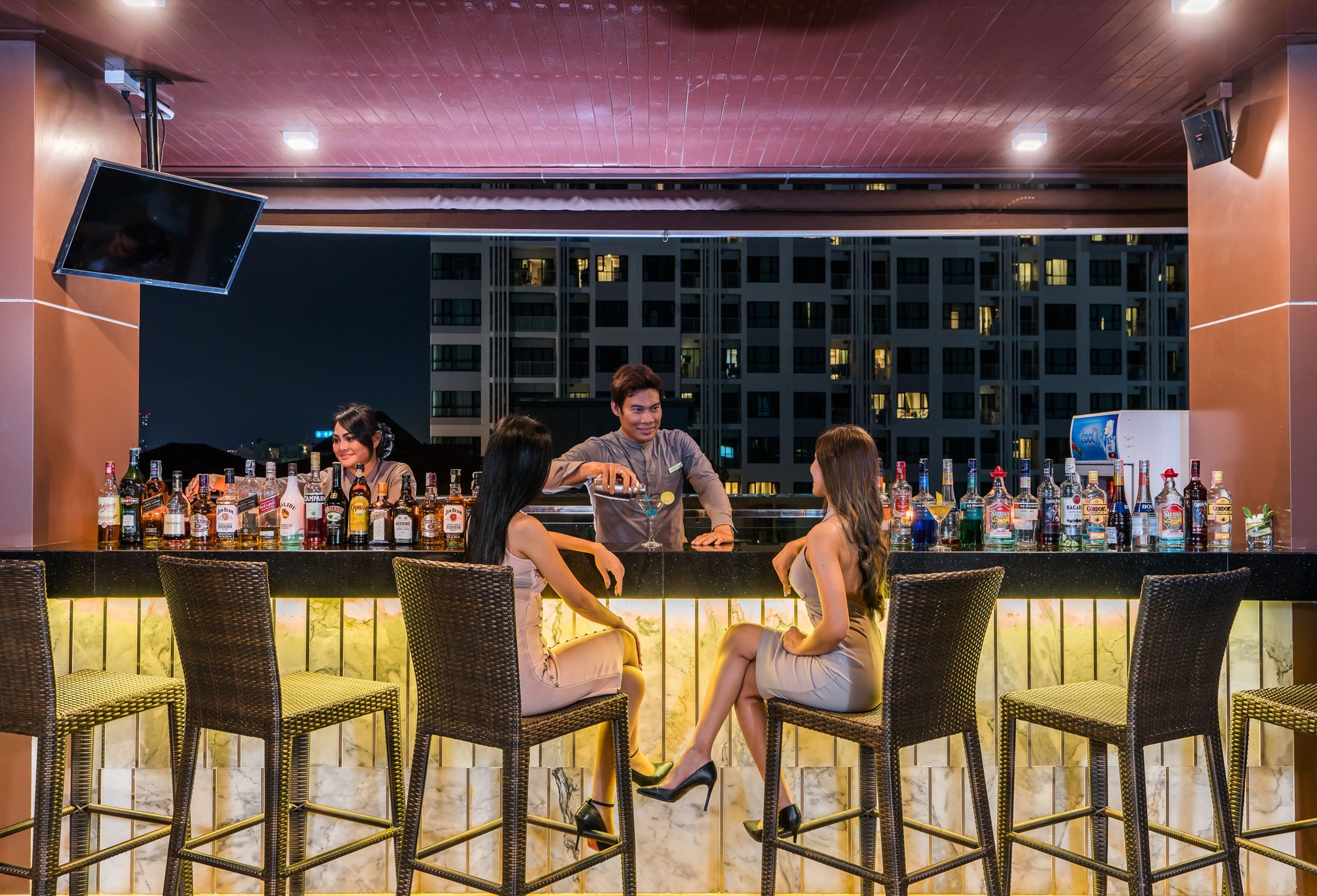 A bar area with wicker chairs, a counter with various bottles, and a cityscape view in the background.