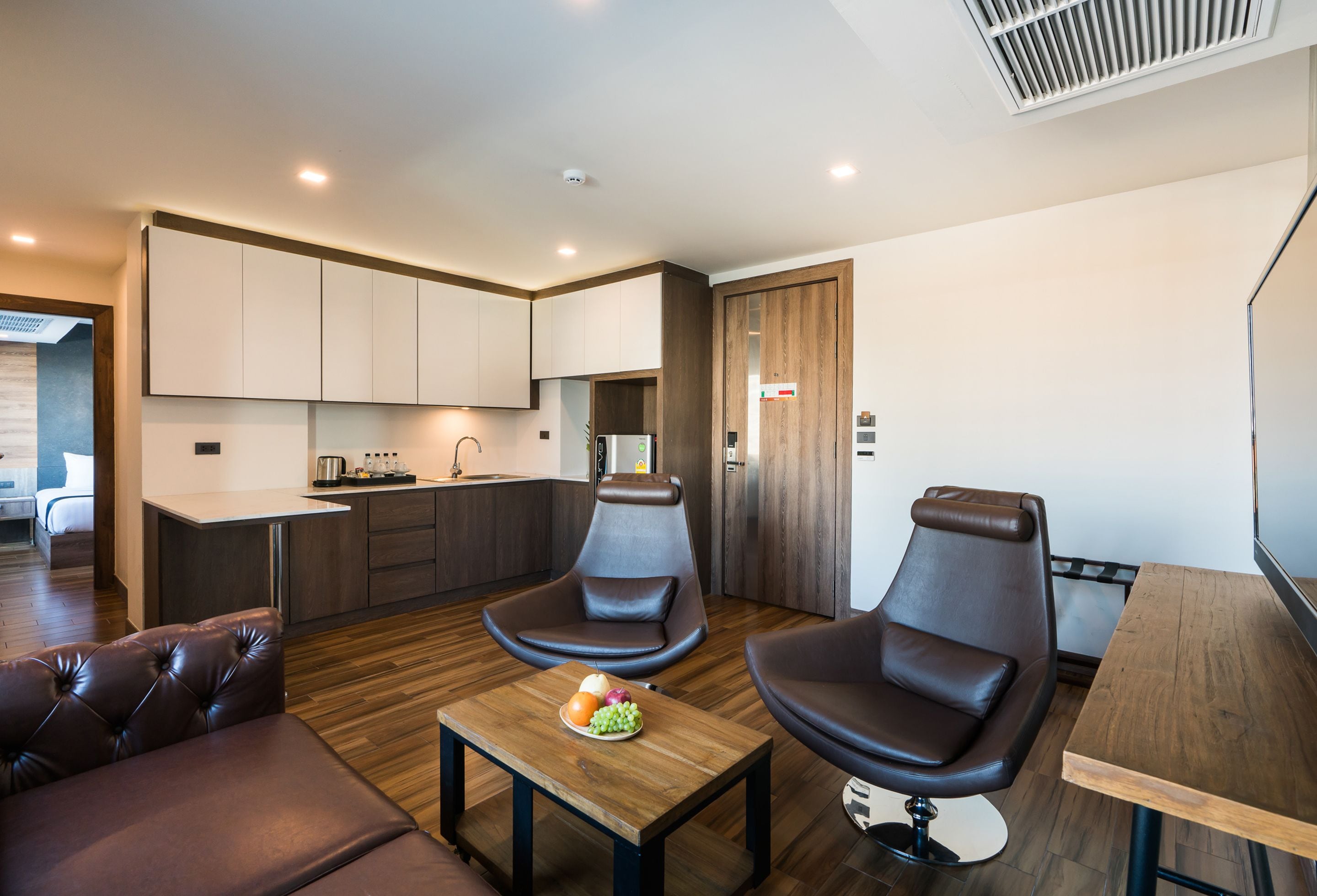 A modern living room with a sofa, two black leather chairs, a wooden coffee table with fruit, and a kitchen in the background.