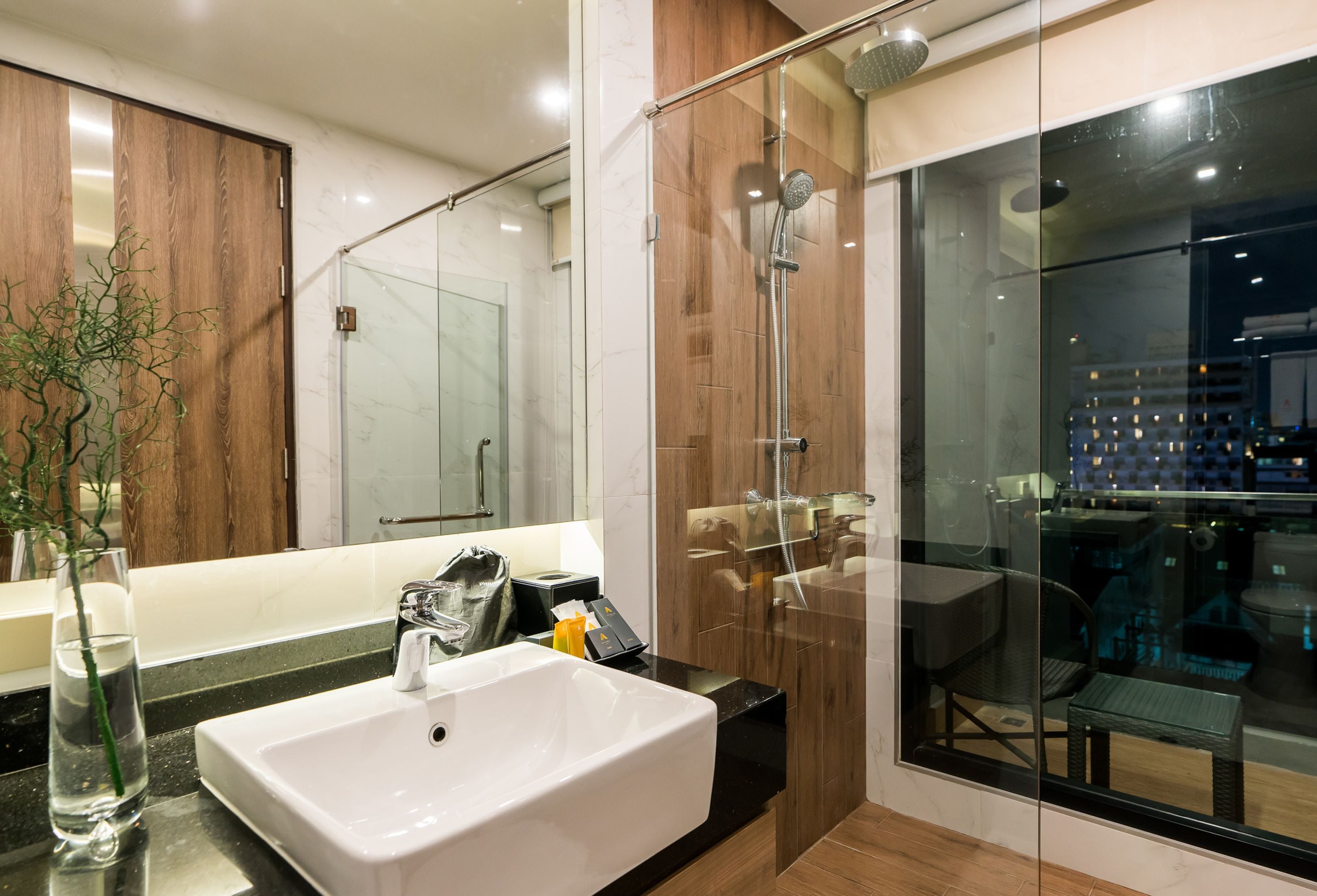 Une salle de bain moderne avec une douche à parois de verre, un grand miroir et un lavabo agrémenté d'un vase de plantes vertes.