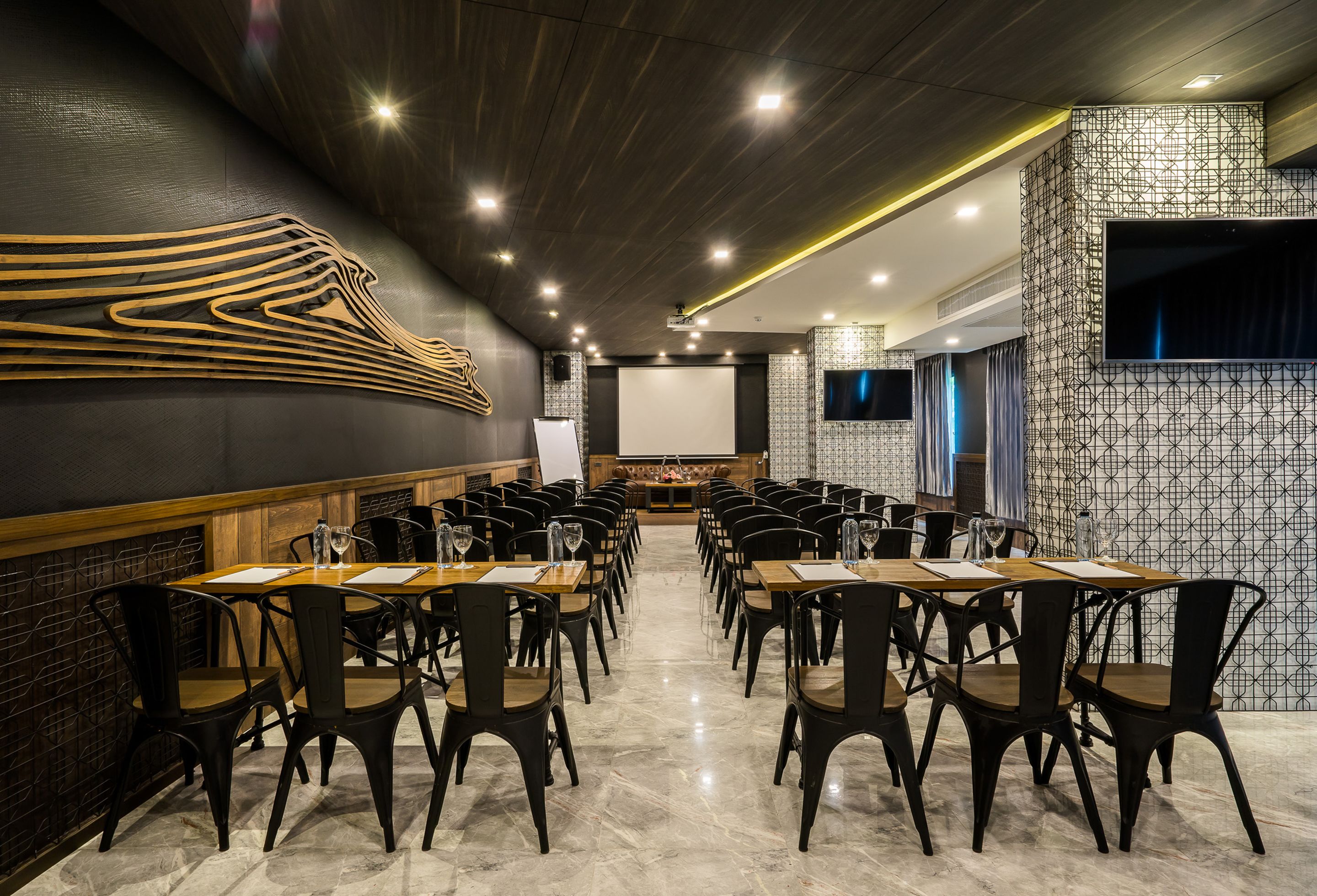 A conference room with a long table, chairs, a projector screen, and a patterned wall.