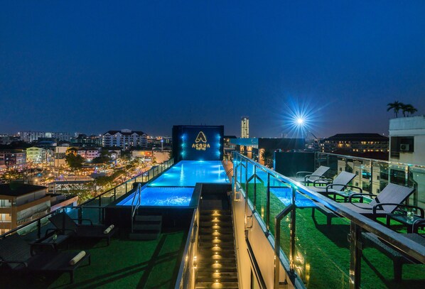 Une piscine sur le toit offrant une vue sur la ville la nuit, un panneau avec le mot « ACQUA », et des chaises longues disposées le long du bord de la piscine.