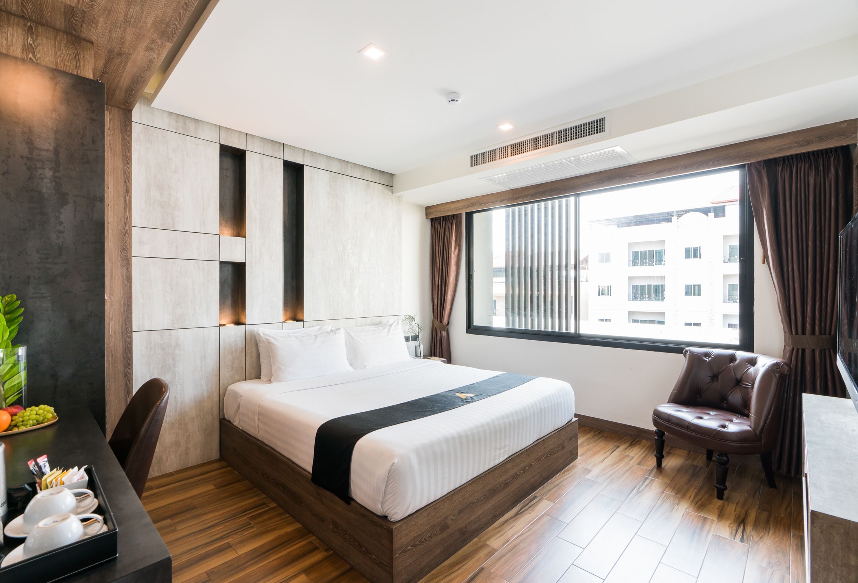 Ein modernes Hotelzimmer mit einem großen Bett, einem Holz-Kopfteil, einem Schreibtisch mit Stuhl und einem großen Fenster mit Vorhängen.