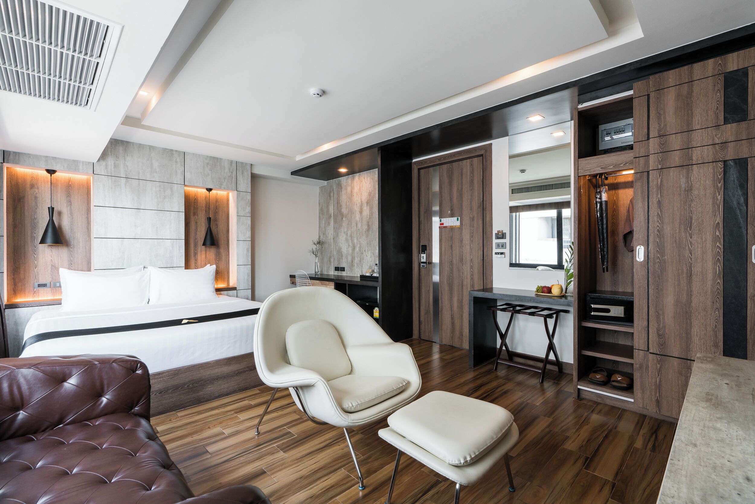 A modern hotel room with a large bed, a white armchair, a brown leather sofa, and a wooden floor.