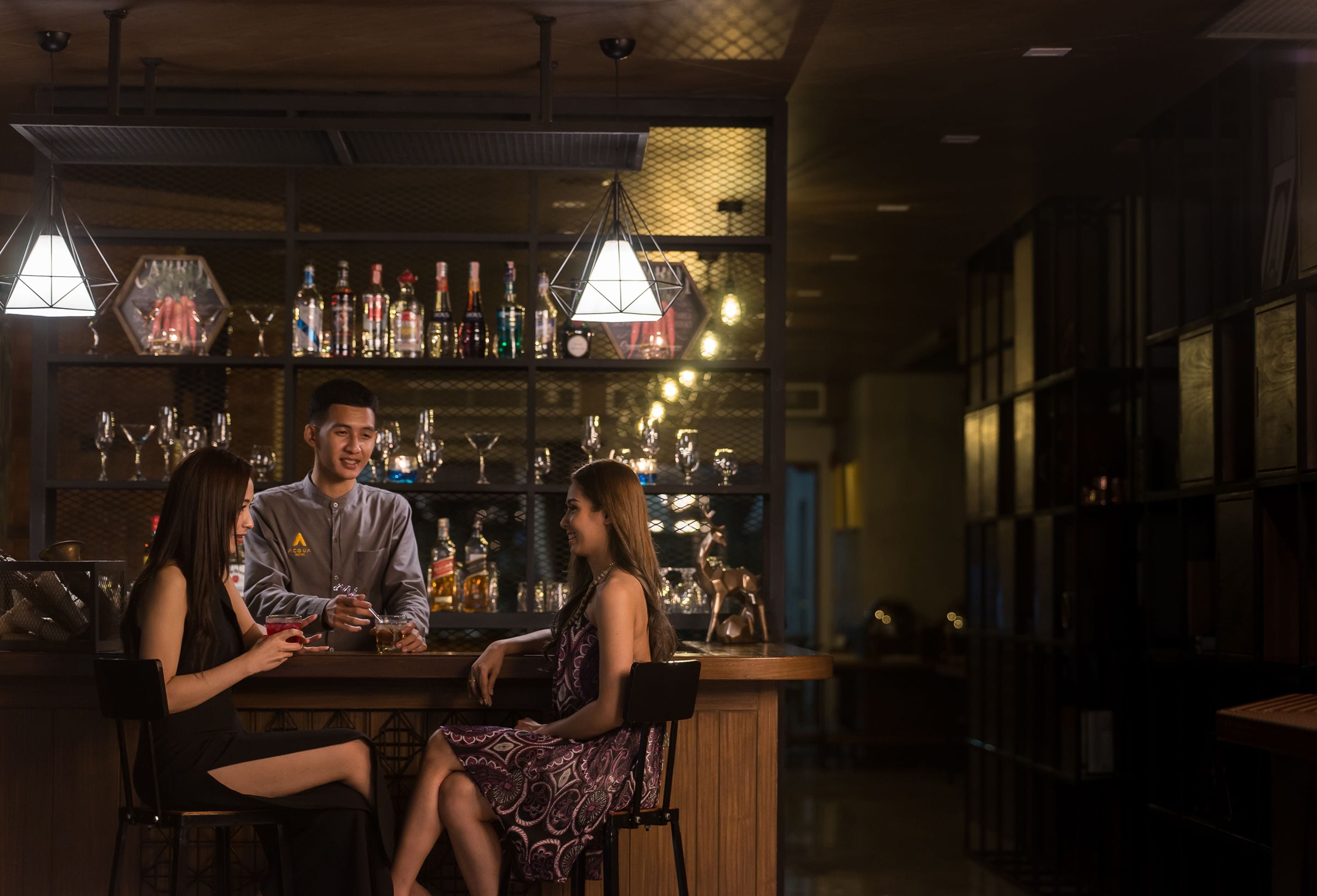 A bar area with a bartender, two seated guests, and a well-stocked bar counter.