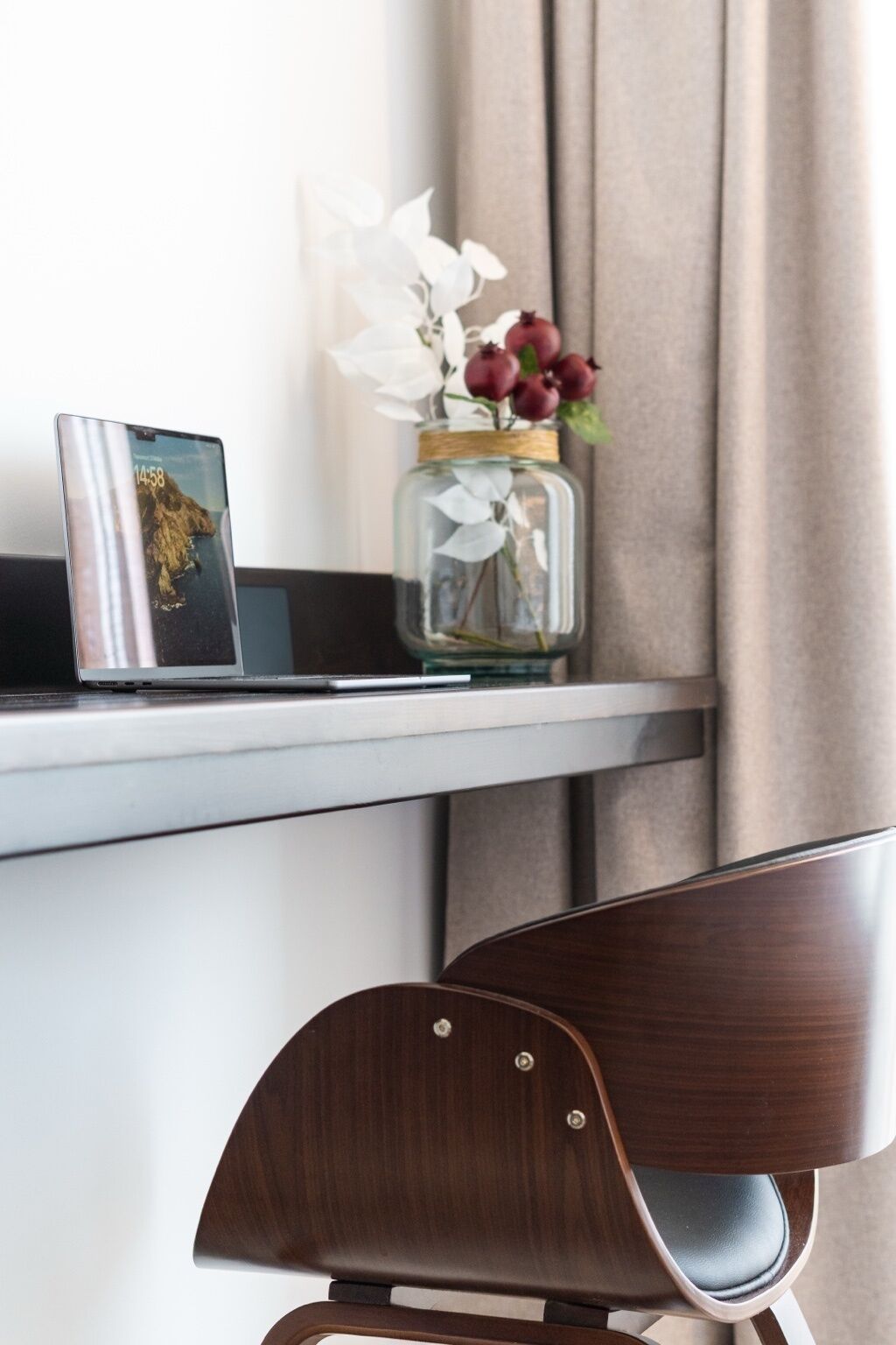 Una computadora portátil sobre un escritorio blanco, un florero con flores y una silla de madera.