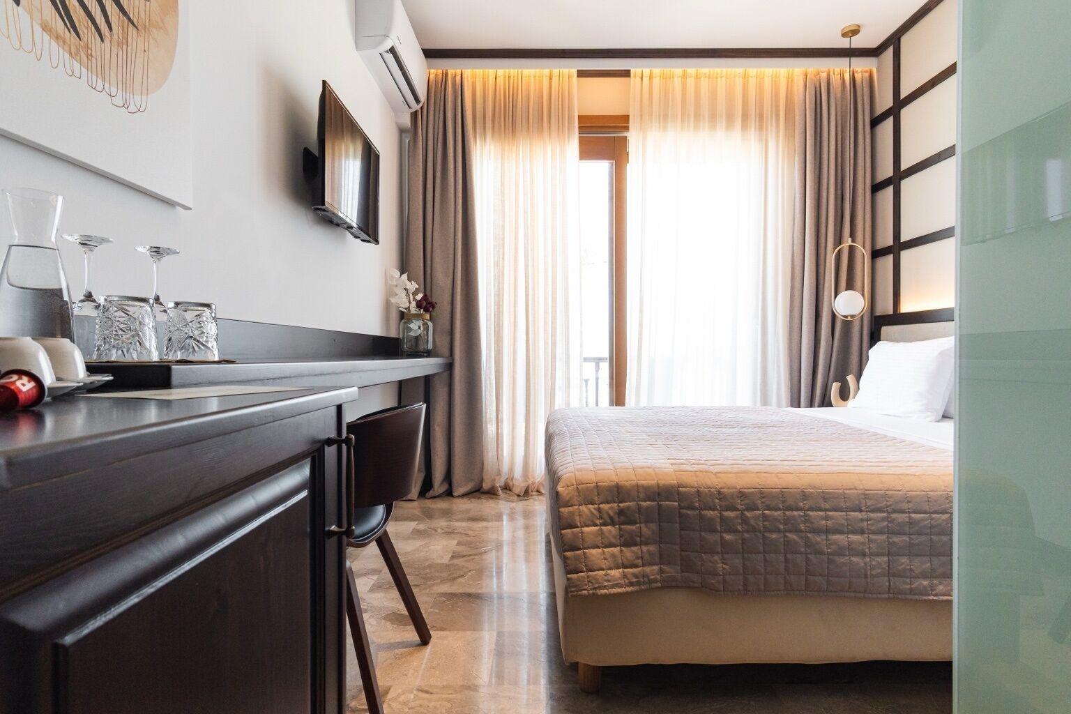 A modern hotel room with a bed, a desk with chairs, a TV, and a large window with curtains.