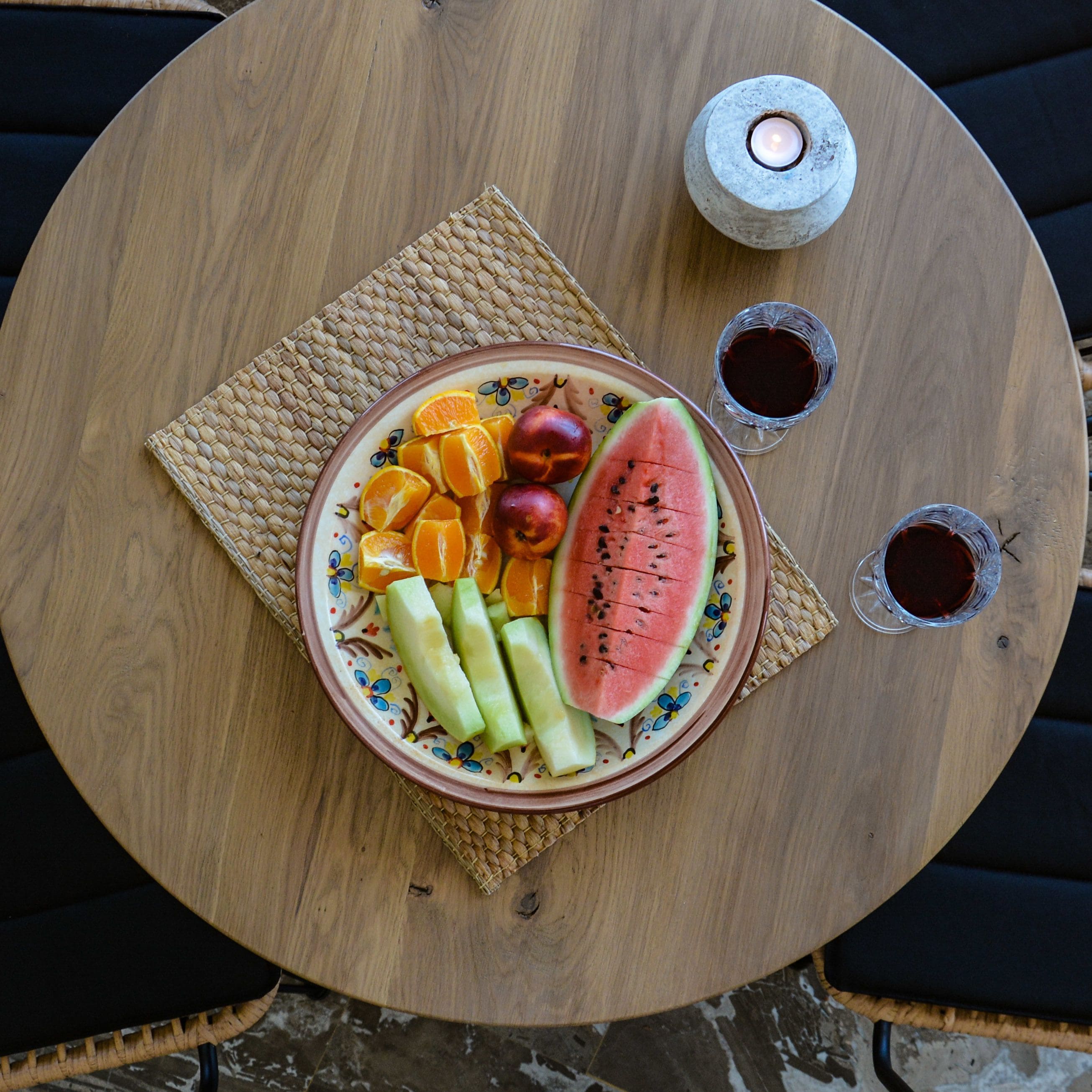 A round wooden table with a woven placemat, a plate of fruit, and two glasses of dark liquid.
