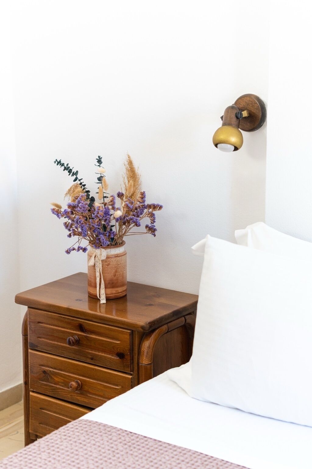 Una mesita de noche de madera con un jarrón de flores secas, una lámpara de pared y una cama con un almohadón blanco.