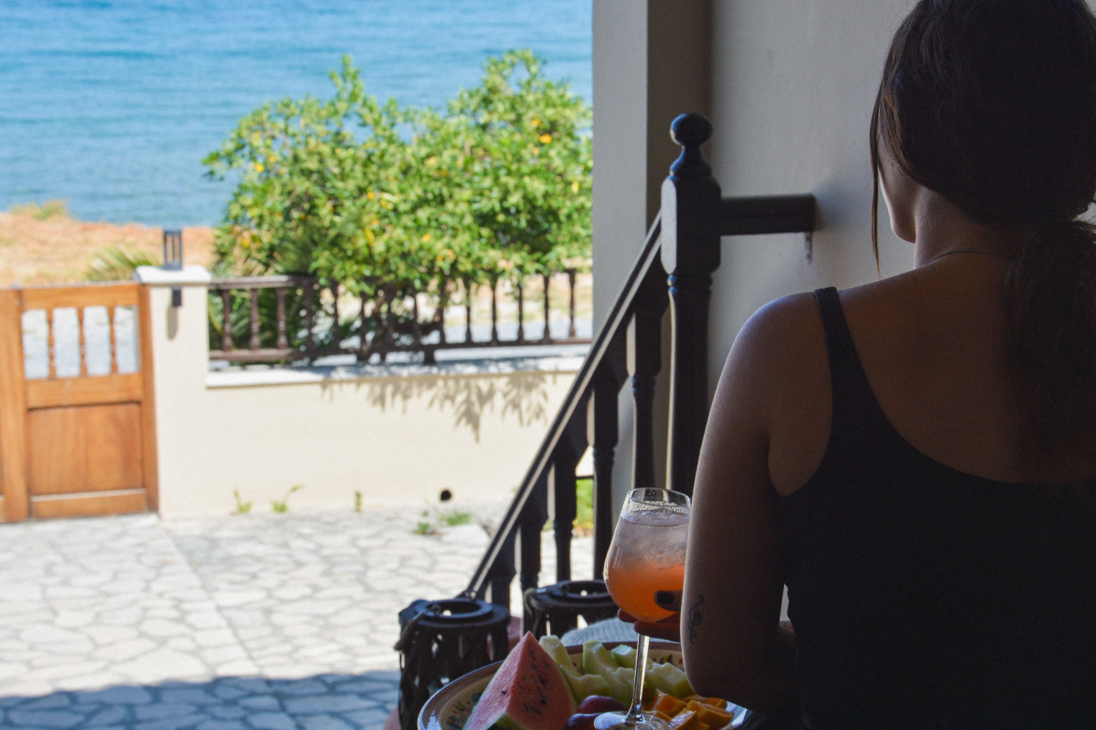 Une personne est assise à une table, devant une boisson et des fruits, et elle a devant elle une vue sur une zone côtière, avec une barrière en bois et de la végétation.
