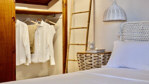 A bedroom with a bed, a wardrobe with hanging clothes, a bedside table with a book and a candle, and a woven side table. - Sunrise Hotel (Samos)
