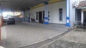 A covered parking area with a car, a building with blue and white columns, and a staircase leading to the entrance. - Losmen Ibu Hj. Tarjo (Palembang)